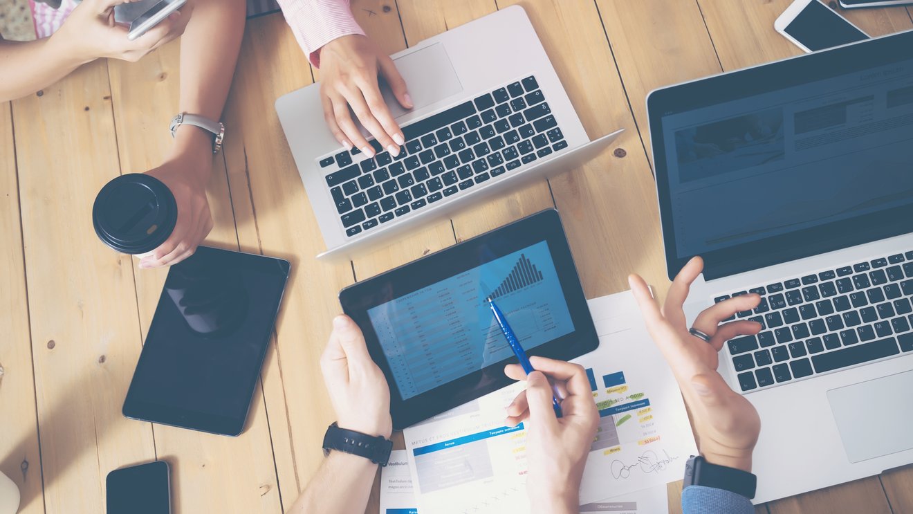 Generisches B&uuml;ro, mehrere H&auml;nde die &uuml;ber einen Tisch greifen Laptops und Tablets auf dem tisch Kachel Vorschau