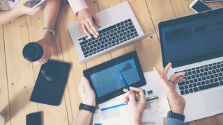 Generisches B&uuml;ro, mehrere H&auml;nde die &uuml;ber einen Tisch greifen Laptops und Tablets auf dem tisch Kachel Vorschau
